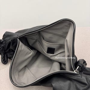 May include: Black leather handbag with a silver zipper and a gray interior. The bag is open, revealing a spacious compartment and a small pocket. The bag has a soft, slouchy design and a knotted detail on the side.