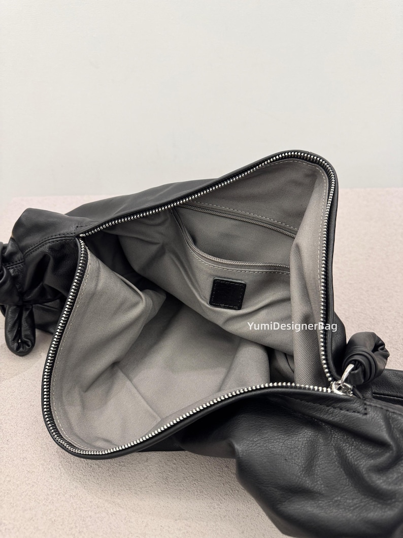 May include: Black leather handbag with a silver zipper and a gray interior. The bag is open, revealing a spacious compartment and a small pocket. The bag has a soft, slouchy design and a knotted detail on the side.