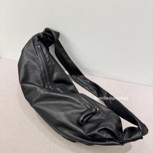 May include: A black leather shoulder bag with a long strap and a zipper closure. The bag is made of a soft, pliable material and has a knotted detail on the strap. The bag is designed to be worn over the shoulder.