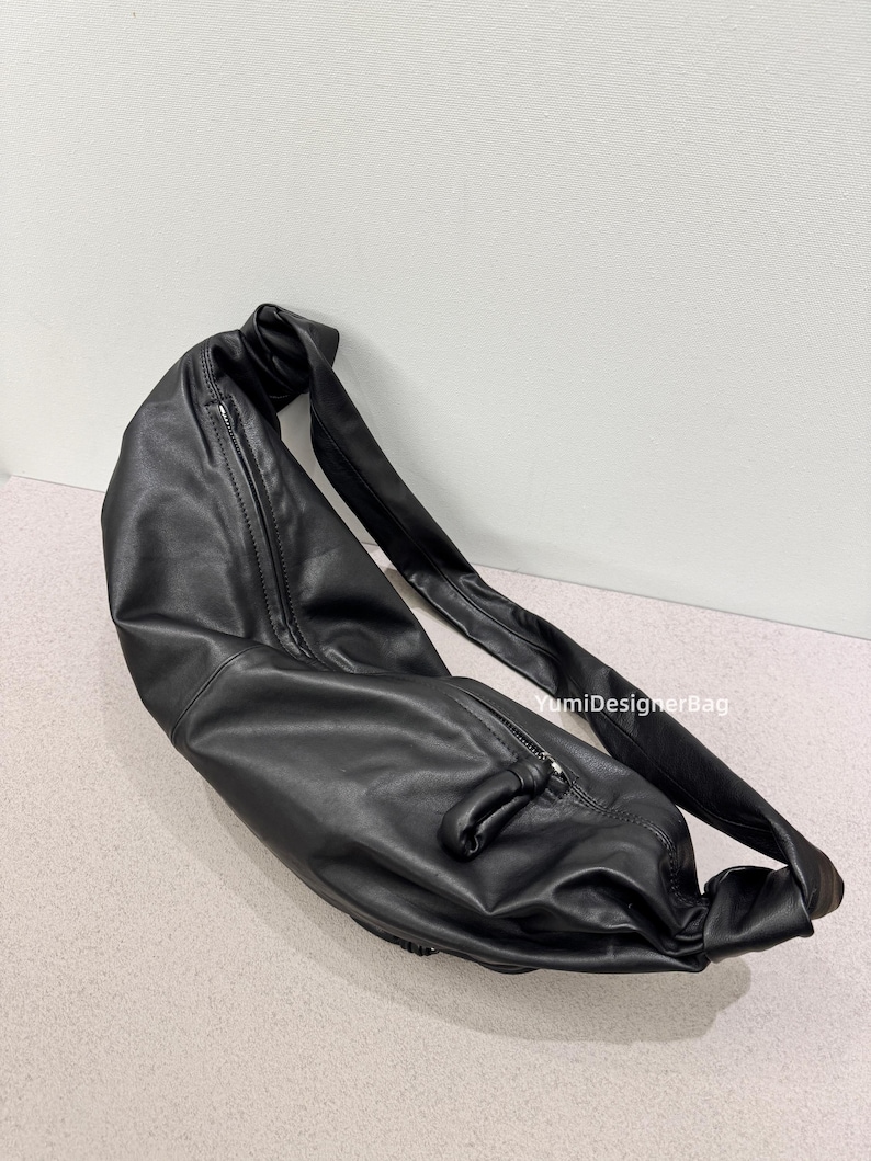 May include: A black leather shoulder bag with a long strap and a zipper closure. The bag is made of a soft, pliable material and has a knotted detail on the strap. The bag is designed to be worn over the shoulder.