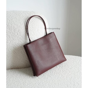 May include: A deep burgundy square-shaped handbag with two thin, rounded top handles. The bag features a smooth, matte finish and a clean, structured silhouette. The text YumiDesignerBag is printed in small black letters on the upper right side.