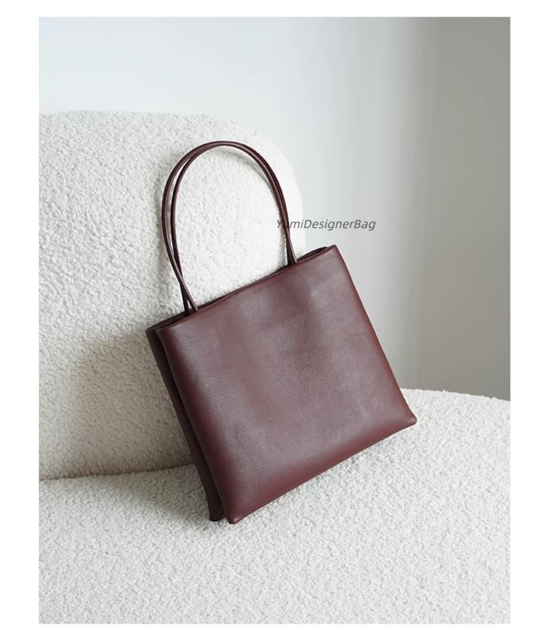 May include: A deep burgundy square-shaped handbag with two thin, rounded top handles. The bag features a smooth, matte finish and a clean, structured silhouette. The text YumiDesignerBag is printed in small black letters on the upper right side.