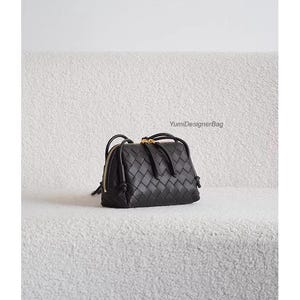 K&ouml;nnte beinhalten: Schwarze Handtasche aus geflochtenem Leder mit goldenem Rei&szlig;verschluss und Griffen. Die Tasche hat eine rechteckige Form und ein geflochtenes Muster. Der Text "YumiDesignerBag" ist im Hintergrund sichtbar.