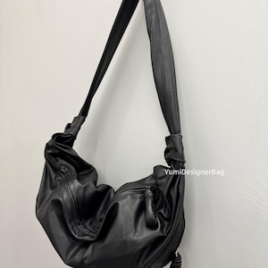 May include: Black leather shoulder bag with a soft, slouchy design. The bag features a long, adjustable strap and a zippered pocket on the front. The bag is hanging on a wooden hook against a white wall.