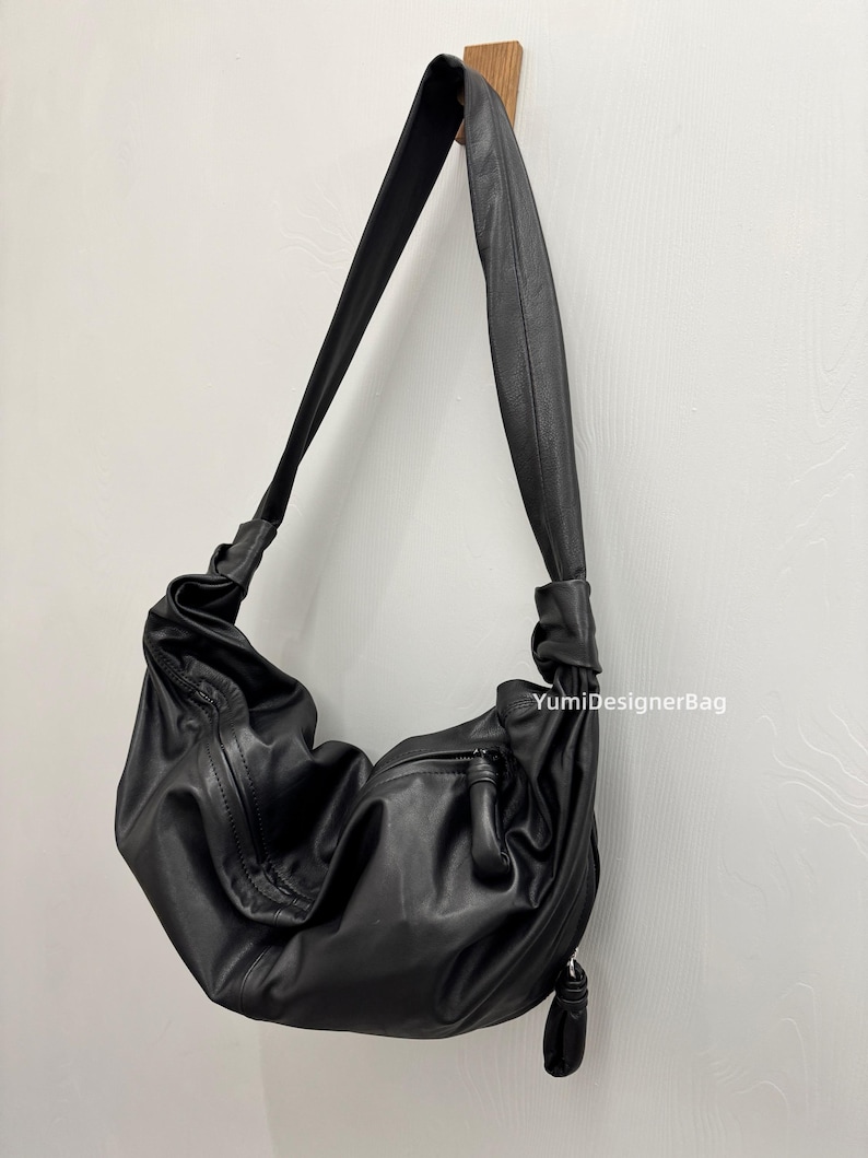 May include: Black leather shoulder bag with a soft, slouchy design. The bag features a long, adjustable strap and a zippered pocket on the front. The bag is hanging on a wooden hook against a white wall.
