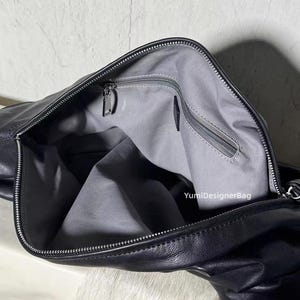 May include: Black leather handbag with a silver zipper closure. The interior is lined with gray fabric and features a zippered pocket. The bag is open, revealing the inside. The brand name "YumiDesignerBag" is visible.