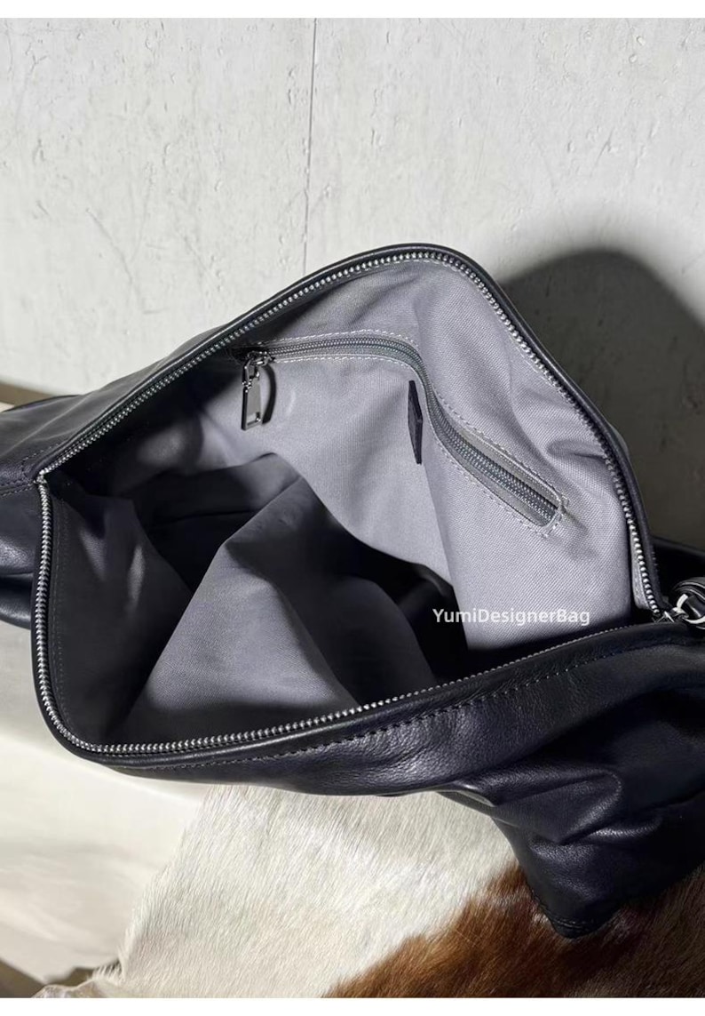 May include: Black leather handbag with a silver zipper closure. The interior is lined with gray fabric and features a zippered pocket. The bag is open, revealing the inside. The brand name "YumiDesignerBag" is visible.