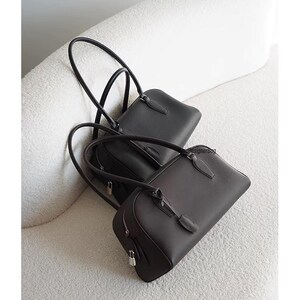 May include: Two leather handbags in black and dark brown rest on a white, textured surface. The bags have long, curved handles and a rectangular shape with a zipper closure. The bags are a classic design.