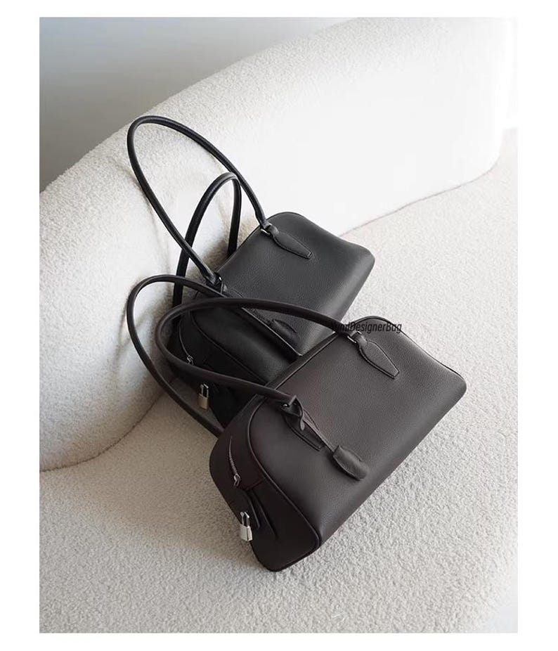May include: Two leather handbags in black and dark brown rest on a white, textured surface. The bags have long, curved handles and a rectangular shape with a zipper closure. The bags are a classic design.
