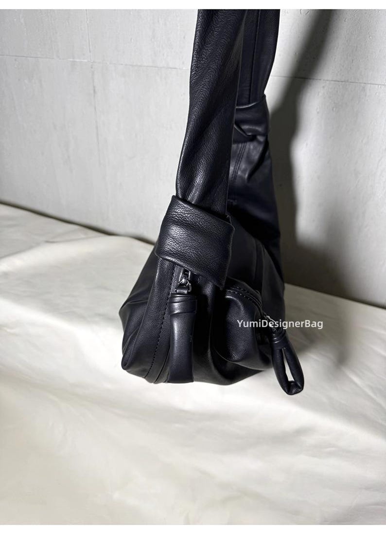 May include: A black leather shoulder bag with a soft, slouchy design. The bag features a zippered pocket, a small exterior pocket, and a knotted handle detail. The bag is made of smooth leather and has a minimalist aesthetic.