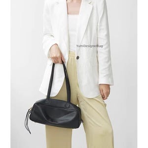 May include: A black leather handbag with long handles is held in front of a person wearing a white blazer and light yellow pants. The bag has a rectangular shape with a zipper closure and a small tassel detail. The text "YumiDesignerBag" is visible.