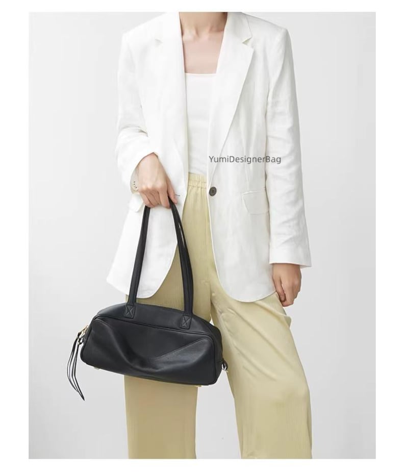 May include: A black leather handbag with long handles is held in front of a person wearing a white blazer and light yellow pants. The bag has a rectangular shape with a zipper closure and a small tassel detail. The text "YumiDesignerBag" is visible.