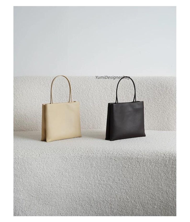 May include: Two rectangular tote bags with thin handles, one in beige and one in black, featuring a smooth finish and a structured silhouette. The text YumiDesignerBag is visible in the center above the black bag.