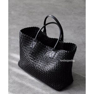 Puede incluir: Bolso tote negro tejido con dos asas redondeadas. El bolso tiene forma rectangular y un patrón tejido. Las asas están unidas con detalles de cuero. El bolso está sobre una superficie gris. El texto "YumiDesignerBag" es visible.