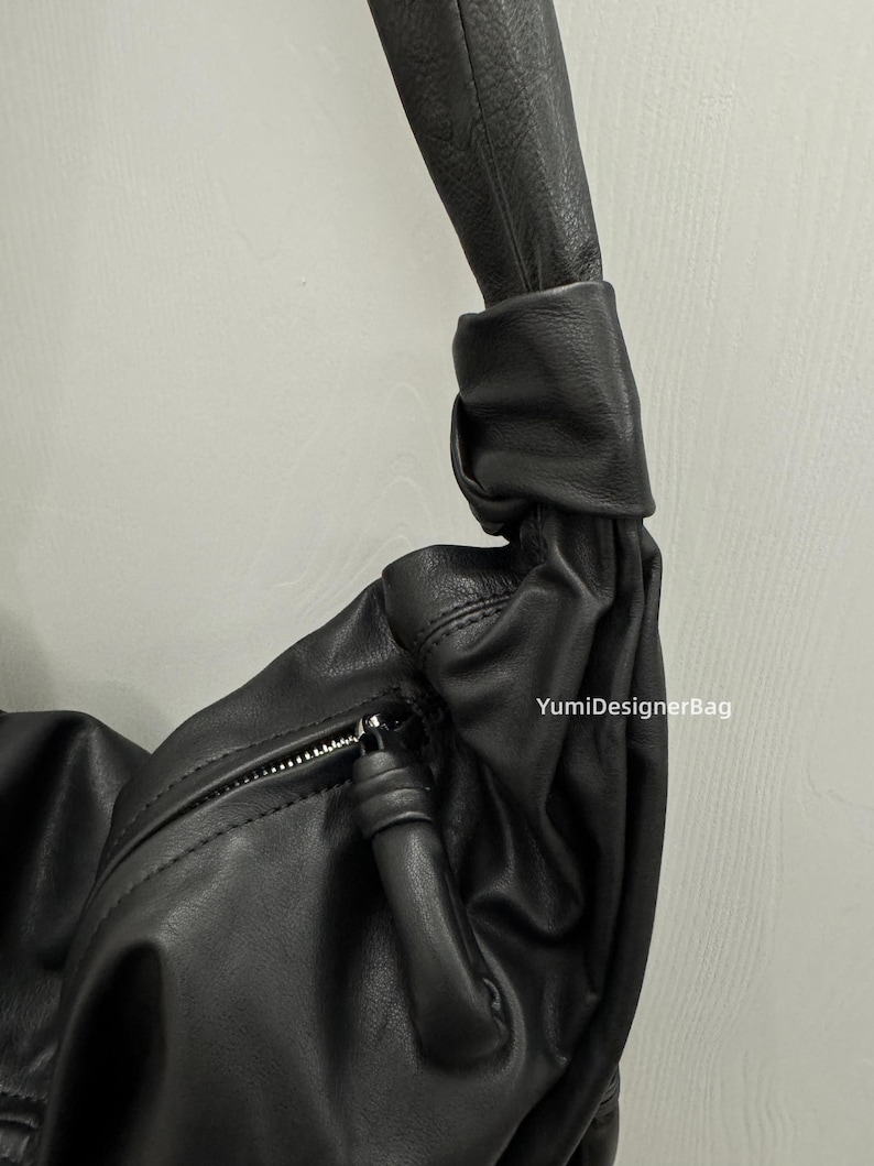 May include: Black leather shoulder bag with a knotted handle. The bag features a zipper closure and a soft, textured appearance. The bag is a dark, solid color and appears to be made of high-quality leather.