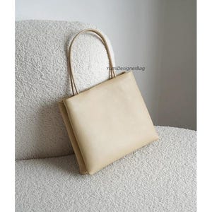May include: A beige square-shaped handbag with two thin matching handles. The bag features a smooth, solid-colored surface and includes the text YumiDesignerBag printed in small black letters on the upper right side of the front panel.