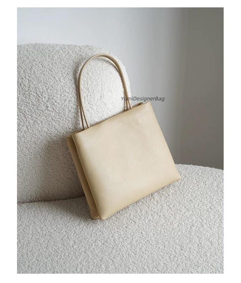 May include: A beige square-shaped handbag with two thin matching handles. The bag features a smooth, solid-colored surface and includes the text YumiDesignerBag printed in small black letters on the upper right side of the front panel.
