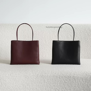 May include: Two square-shaped handbags with thin top handles, one in deep burgundy and one in black. The bags feature a smooth, solid-colored exterior and a clean, structured design. The text YumiDesignerBag is visible between the two bags.