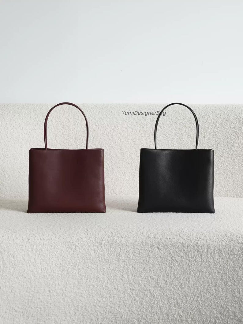 May include: Two square-shaped handbags with thin top handles, one in deep burgundy and one in black. The bags feature a smooth, solid-colored exterior and a clean, structured design. The text YumiDesignerBag is visible between the two bags.