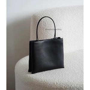 May include: A black square-shaped handbag with a smooth finish and two thin, rounded top handles. The bag features a structured, upright design with a clean silhouette. The text YumiDesignerBag is printed in small black letters on the upper center.