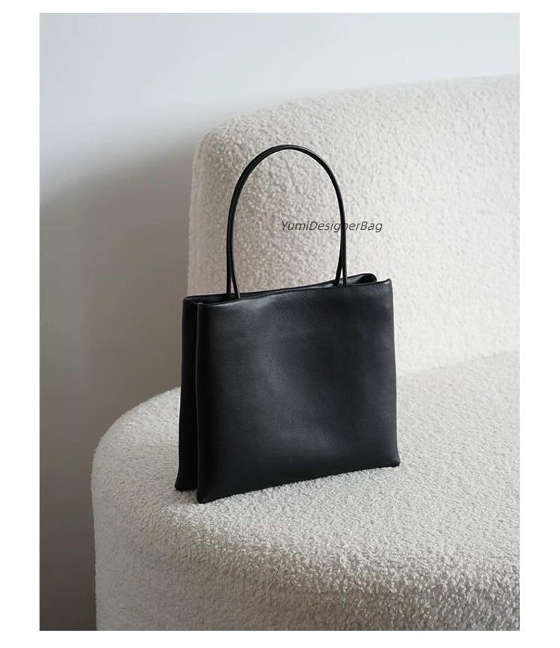 May include: A black square-shaped handbag with a smooth finish and two thin, rounded top handles. The bag features a structured, upright design with a clean silhouette. The text YumiDesignerBag is printed in small black letters on the upper center.