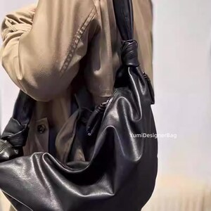 May include: A black leather shoulder bag with a knotted strap. The bag has a soft, slouchy design and is worn over the shoulder. The bag is made of a smooth, matte leather. The text "YumiDesignerBag" is visible on the bag.