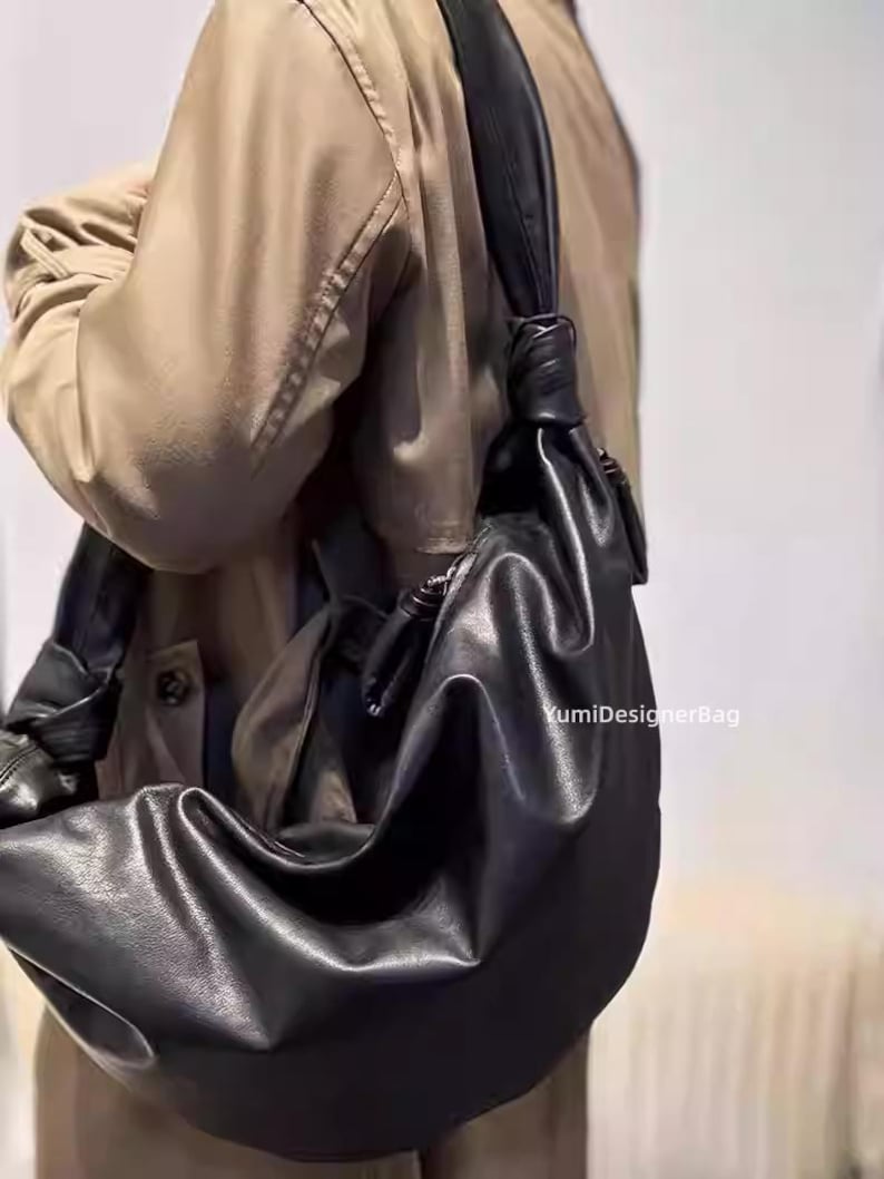 May include: A black leather shoulder bag with a knotted strap. The bag has a soft, slouchy design and is worn over the shoulder. The bag is made of a smooth, matte leather. The text "YumiDesignerBag" is visible on the bag.