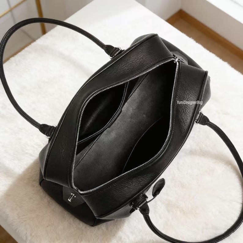 May include: Black leather handbag with silver zippers and handles. The bag is open, revealing the interior compartments. The handles are curved and attached to the top of the bag. The bag is sitting on a white surface.