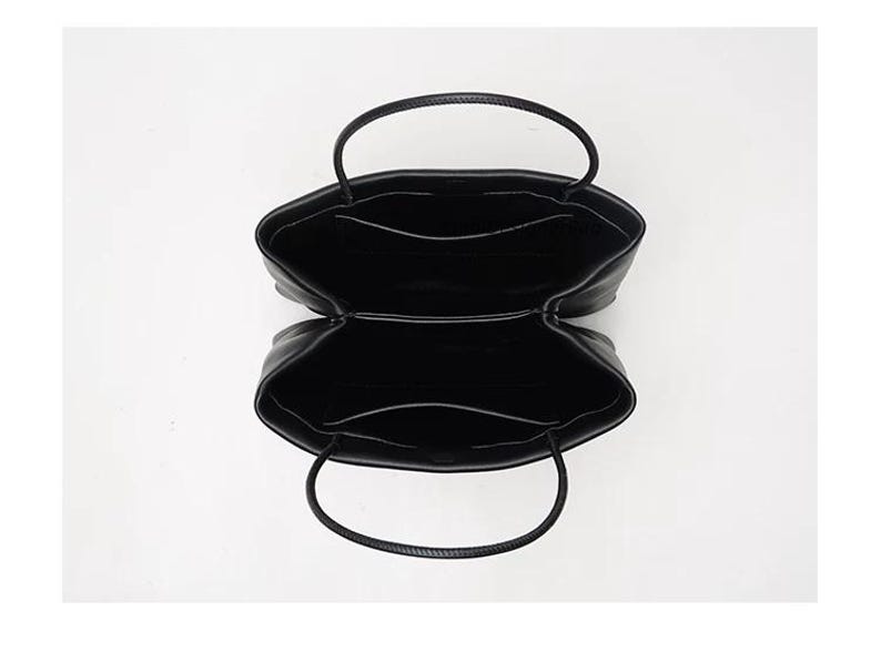May include: A black handbag viewed from directly above, showing an open interior divided into two main compartments. Each compartment features a flat pocket along the inner wall and a thin, rounded handle extending from the top edge.