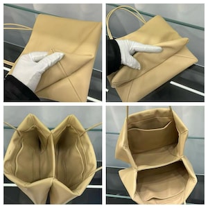 May include: A beige shoulder bag with a smooth finish, featuring thin straps and a structured, multi-compartment interior. The bag has a soft, pliable exterior and includes internal pockets for organization, with a clean, minimalist design throughout.
