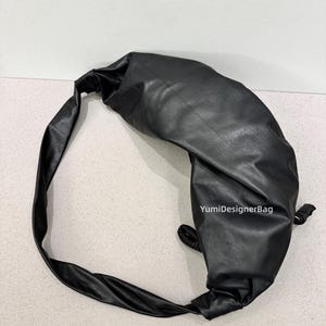 May include: Black leather shoulder bag with a knotted design. The bag has a long strap and a rounded shape. The bag is made of a smooth, shiny material. The text "YumiDesignerBag" is visible on the bag.