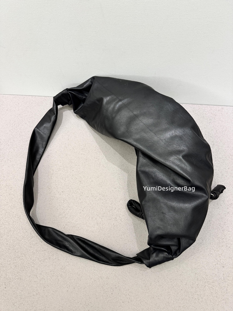 May include: Black leather shoulder bag with a knotted design. The bag has a long strap and a rounded shape. The bag is made of a smooth, shiny material. The text "YumiDesignerBag" is visible on the bag.