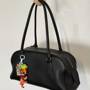 May include: Black leather handbag with long handles and a decorative charm. The bag has a rectangular shape with a zipper closure and a small pocket on the side. The charm features colorful figurines and beads.
