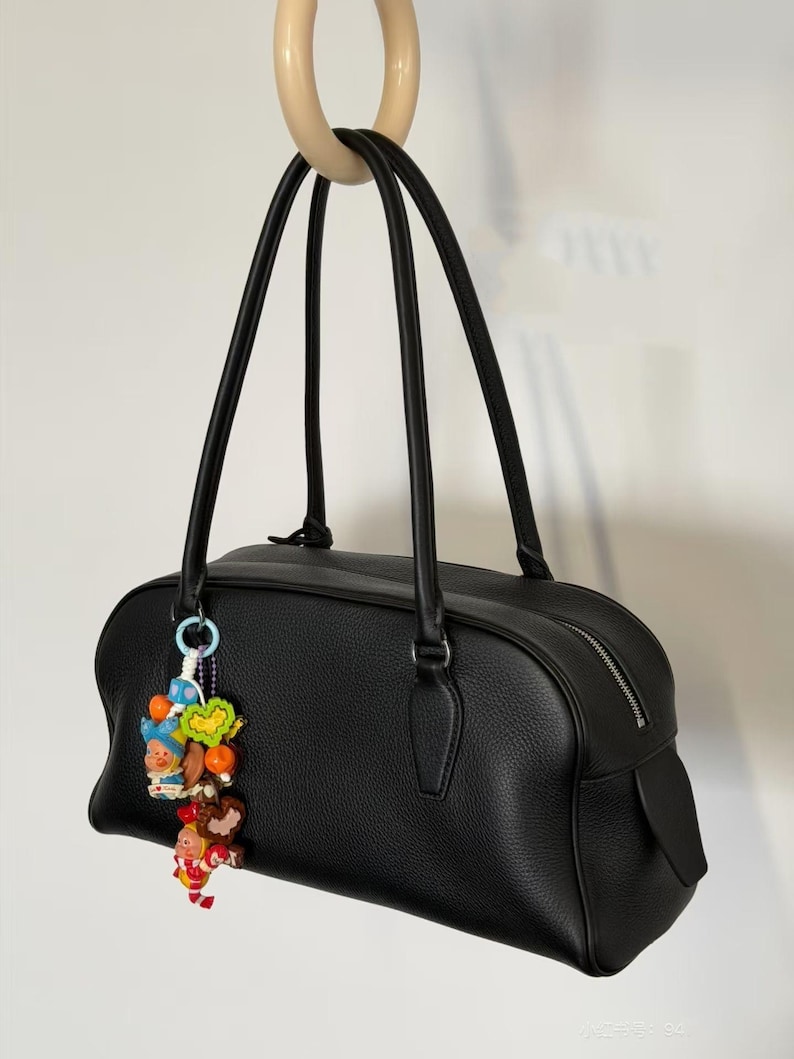May include: Black leather handbag with long handles and a decorative charm. The bag has a rectangular shape with a zipper closure and a small pocket on the side. The charm features colorful figurines and beads.