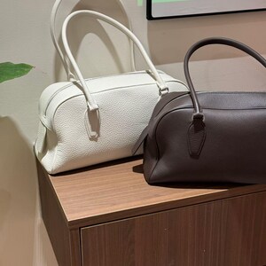 May include: Two leather handbags in the image. One is white, the other is brown. Both have a rectangular shape with rounded corners and top handles. The bags are sitting on a wooden surface.