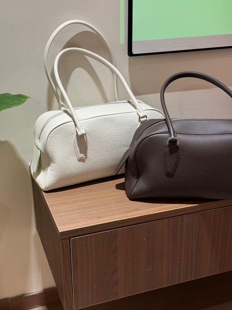 May include: Two leather handbags in the image. One is white, the other is brown. Both have a rectangular shape with rounded corners and top handles. The bags are sitting on a wooden surface.