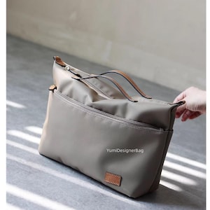 Könnte beinhalten: Eine taupefarbene Tragetasche mit braunen Ledergriffen und -besatz. Die Tasche hat ein Hauptfach mit Reißverschluss und eine Außentasche. Der Text "YumiDesignerBag" ist sichtbar. Die Tasche ist aus einem glatten, strapazierfähigen Material.