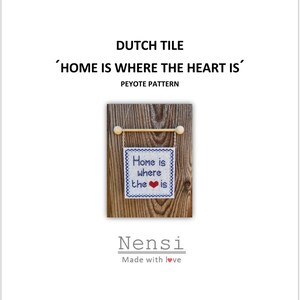 Op de afbeelding: Een klein, vierkant, geborduurd bord met de tekst "Home is where the heart is" en een rood hart. Het bord is blauw en wit en hangt aan een houten deuvel met houten kralen. De achtergrond is een houten oppervlak.