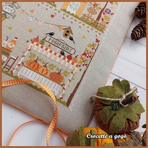 May include: A cross-stitch embroidery of a fall harvest scene with a pumpkin stand, a birdhouse, and a sign that reads "Pumpkins". The embroidery is framed with a light brown border and is surrounded by orange and white checkered ribbon.