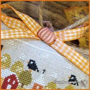 Op de afbeelding: Close-up van een kruissteekontwerp met een herfstthema. Het ontwerp toont een rode schuur, bomen en zwarte vogels. Een oranje en witte ruitjeslint en een pompoenvormige knoop sieren het stuk. De tekst "Croccette a gog&ograve;" is zichtbaar.
