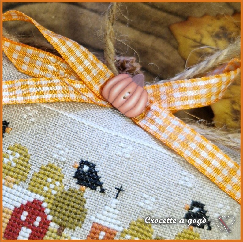 Op de afbeelding: Close-up van een kruissteekontwerp met een herfstthema. Het ontwerp toont een rode schuur, bomen en zwarte vogels. Een oranje en witte ruitjeslint en een pompoenvormige knoop sieren het stuk. De tekst "Croccette a gog&ograve;" is zichtbaar.