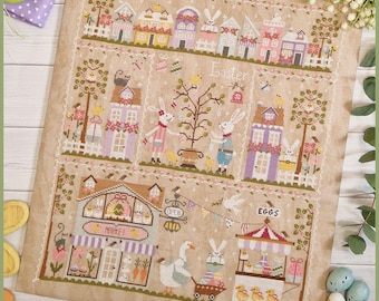 May include: A cross-stitch pattern featuring a charming Easter village scene with pastel-colored houses, a market, an egg stand, and a bunny rabbit. The pattern includes the text "Easter" and "Crocette a gogo".