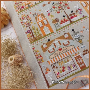 May include: A cross-stitch pattern featuring a charming autumn scene with houses, trees, and a fruit stand. The text "Fresh Fruit" is displayed above the stand, and the words "Crocette a gogo" are at the bottom of the design.
