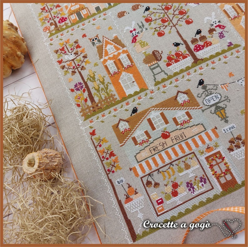 May include: A cross-stitch pattern featuring a charming autumn scene with houses, trees, and a fruit stand. The text "Fresh Fruit" is displayed above the stand, and the words "Crocette a gogo" are at the bottom of the design.