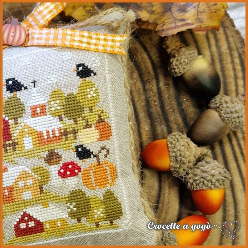 Op de afbeelding: Een kruissteekontwerp met een herfsttafereel met een kerk, pompoenen en bomen. Het ontwerp staat op een beige stof, geaccentueerd met een decoratief oranje en wit geruit lint en een kleine pompoenknop. Eikels zijn gerangschikt op een houten oppervlak.