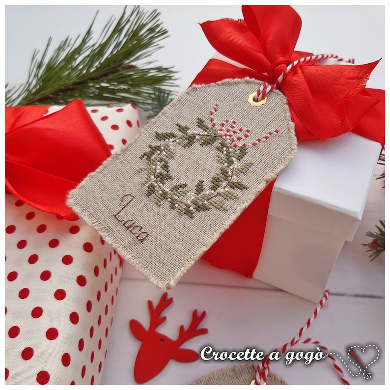 Puede incluir: Una caja de regalo blanca con una cinta roja y una etiqueta de regalo bordada a mano con una corona verde y el nombre "Luca". La etiqueta est&aacute; unida a la caja con un cordel rojo y blanco.