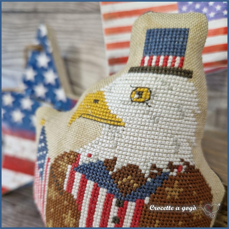 May include: A patriotic, hand-stitched eagle ornament with a top hat, featuring the American flag's colors. The eagle has a yellow beak and is adorned with red, white, and blue details. A star-shaped ornament and a flag are in the background.