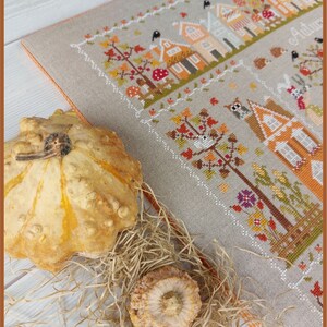 May include: A cross-stitch embroidery of a fall village scene with houses, trees, and birds. The embroidery is framed and displayed on a table with gourds and straw.