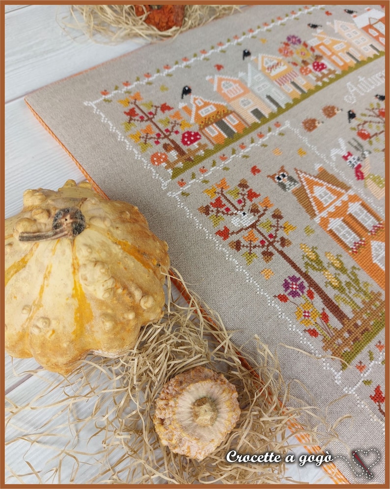 May include: A cross-stitch embroidery of a fall village scene with houses, trees, and birds. The embroidery is framed and displayed on a table with gourds and straw.