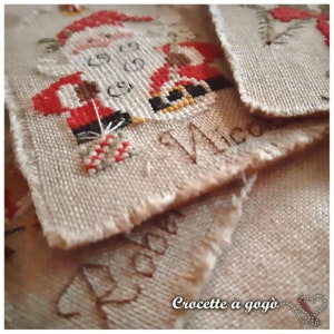 Puede incluir: Primer plano de un adorno de tela beige con un dise&ntilde;o de punto de cruz de Pap&aacute; Noel sosteniendo un bast&oacute;n de caramelo. El adorno est&aacute; etiquetado como "Made" y "Crocette a gog&ograve;".
